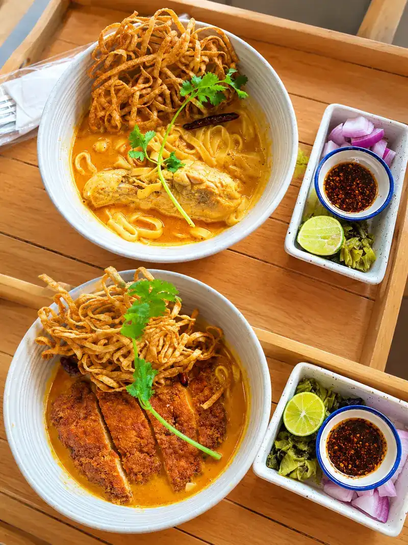 Two bowls of khao soi, one with a chicken drumstick and one with tonkatsu, with shallots, pickled greens and lime on the side
