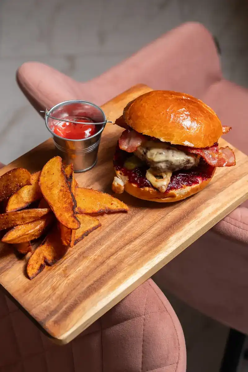 A bacon burger with potato wedges and a cup of ketchup on a wooden board