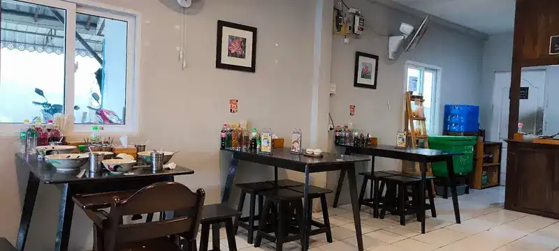 The dining room: dark wooden tables and stools, condiments on the tables, framed pictures and wall fans