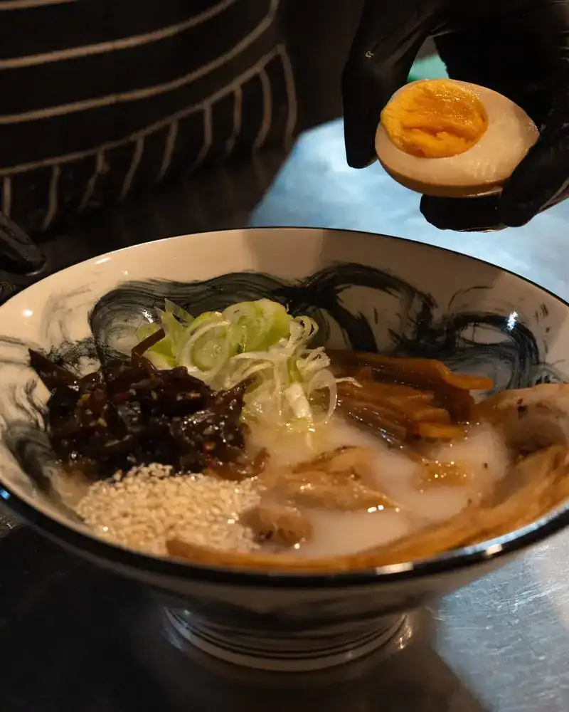 A gloved hand sets half a soft egg onto a finished bowl of ramen