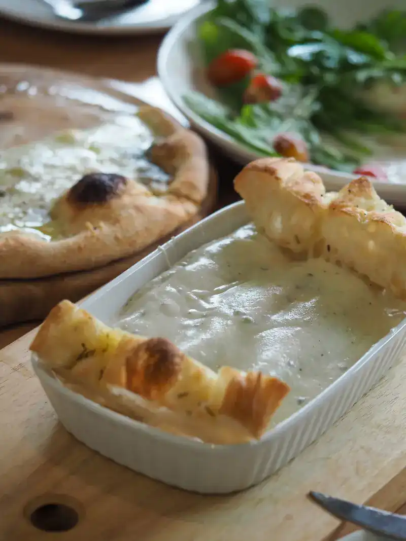 Baked bread sticks resting on a dish of creamy sauce, a pizza behind