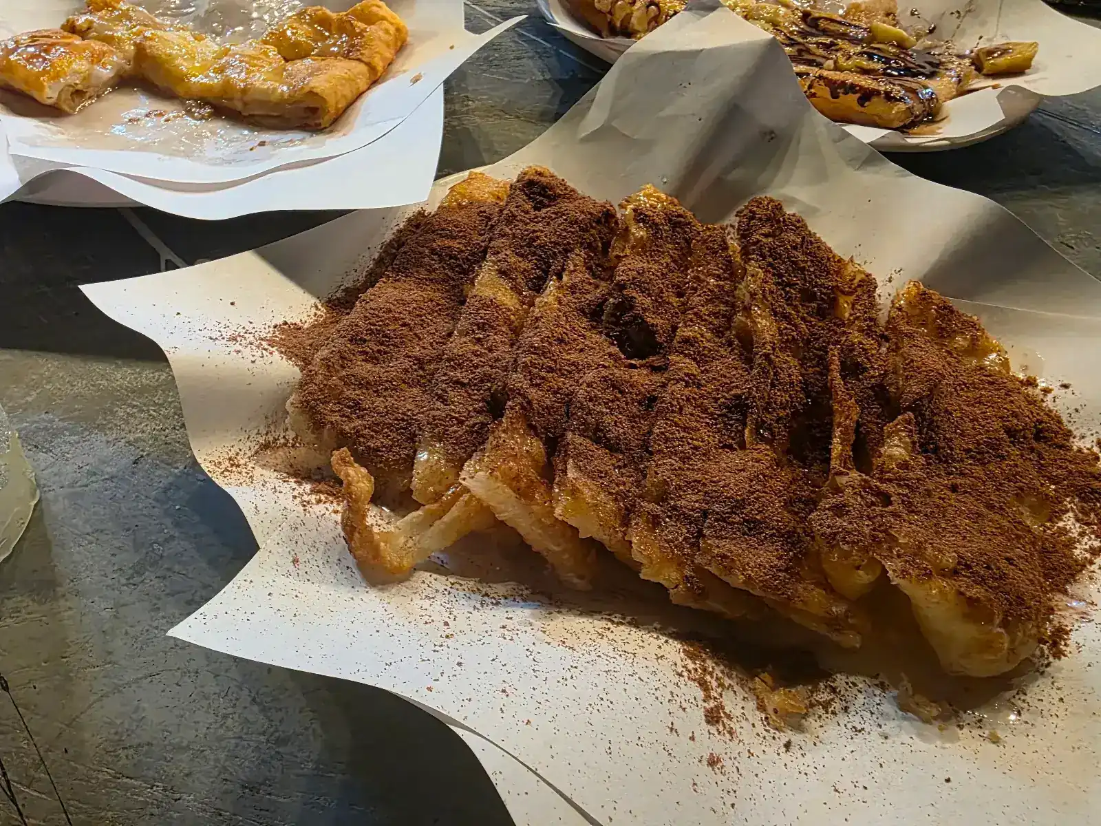 Crispy roti cut into strips and dusted with chocolate powder, on white paper, with more roti behind