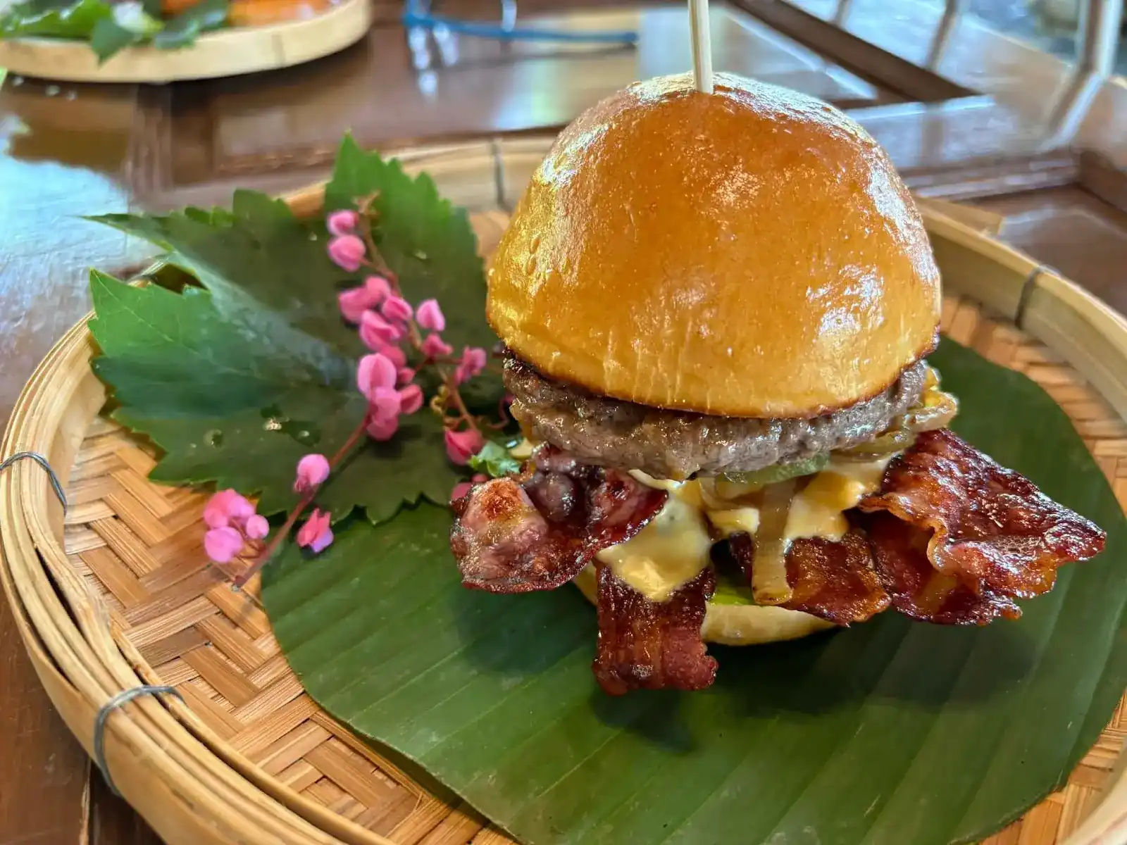 A burger with bacon and melted cheese on a banana leaf in a woven bamboo tray, with pink flowers beside it