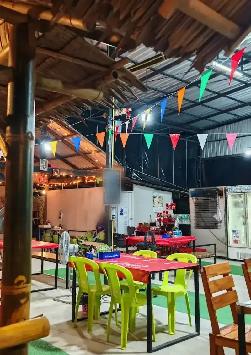 The open-sided dining room at night, with wooden tables, green chairs and coloured bunting under a bamboo and thatch roof / บรรยากาศร้านตอนค่ำ โต๊ะไม้ เก้าอี้สีเขียว และธงราวหลากสี ใต้หลังคาแบบกระท่อมไม้ไผ่