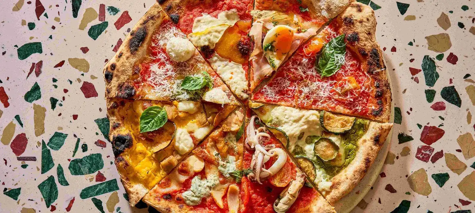 A wood-fired pizza cut into slices, each slice with a different topping, on a terrazzo table