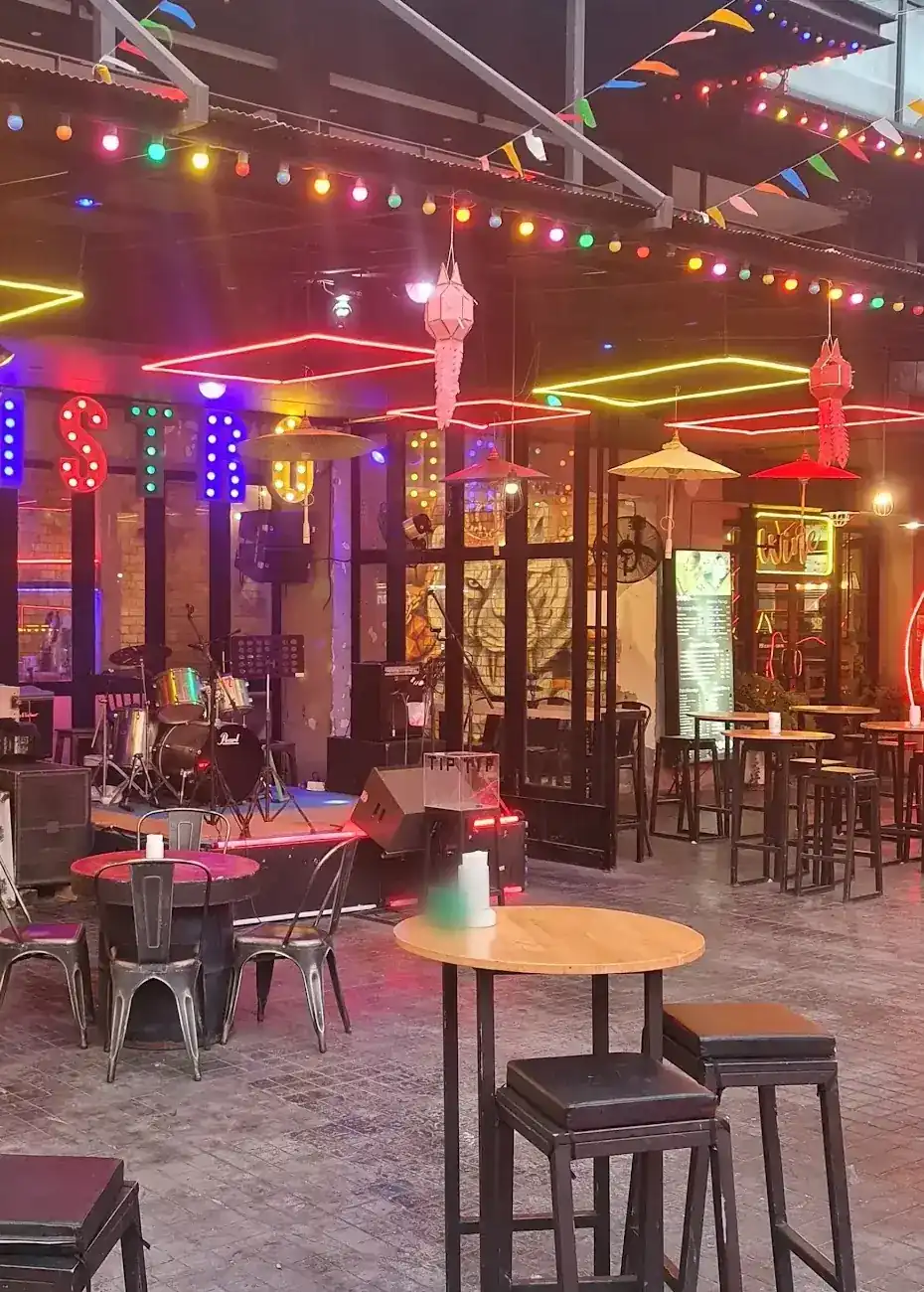 The open-air square at dusk: high tables, a small stage with a drum kit, neon frames and coloured bulbs / ลานกลางแจ้งยามพลบค่ำ โต๊ะสูง เวทีเล็กกับกลองชุด กรอบไฟนีออน และหลอดไฟหลากสี