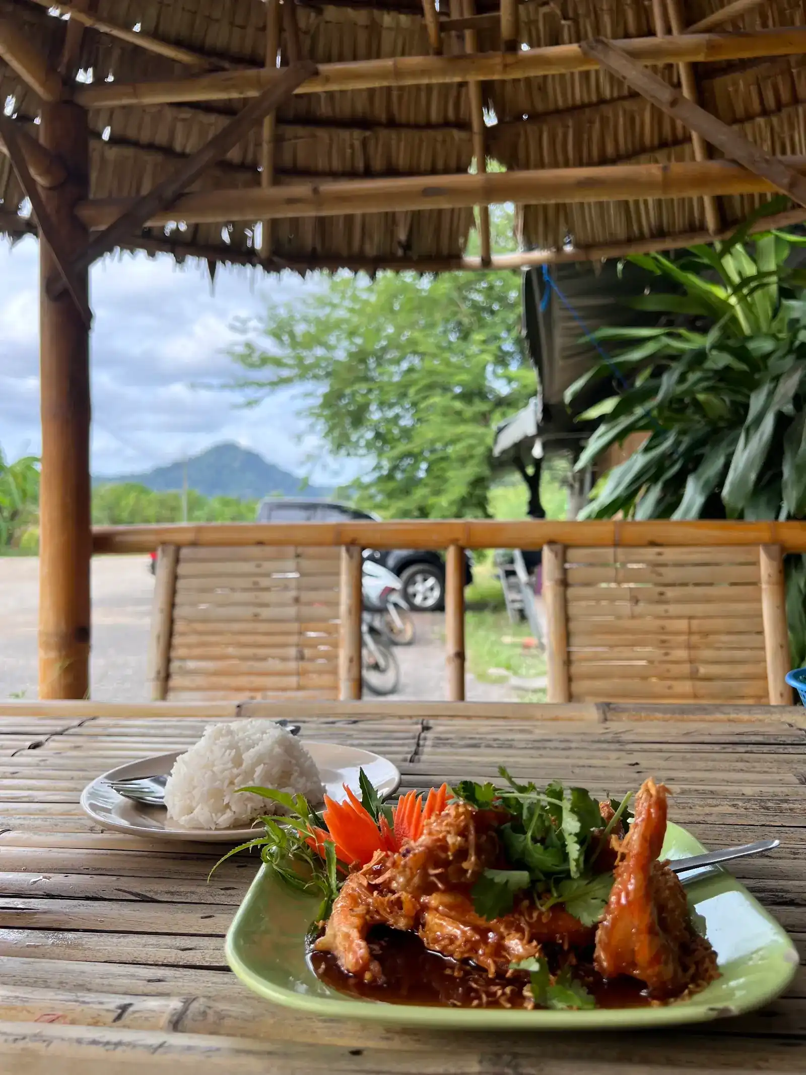 Crispy prawns in a glossy sauce with a plate of rice, on a bamboo table under a thatched roof, a green hill beyond