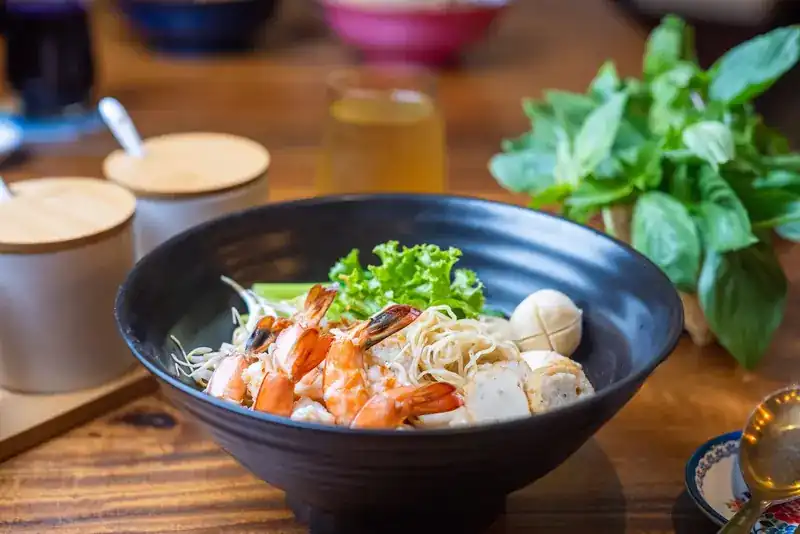 Seafood noodles with prawns and fish balls in a dark bowl, basil behind