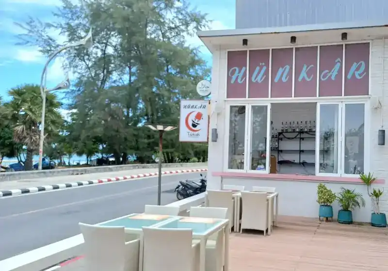 The Hünkâr shopfront on Karon Road: the name across six rose panels, white terrace chairs, and the sea beyond the trees