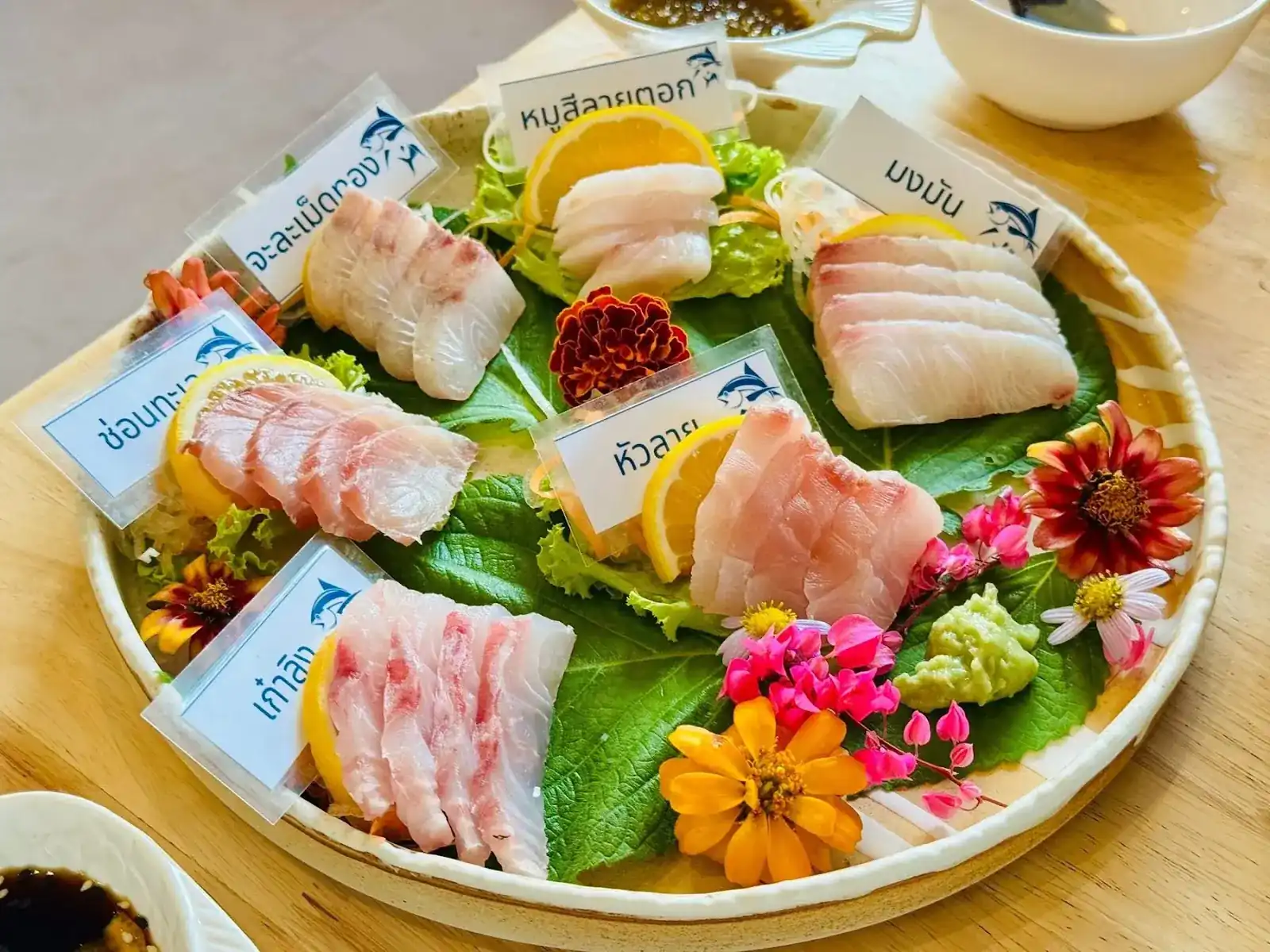 Thai sashimi on a round platter: slices of several fish on leaves with lemon and flowers, each kind named on a small card