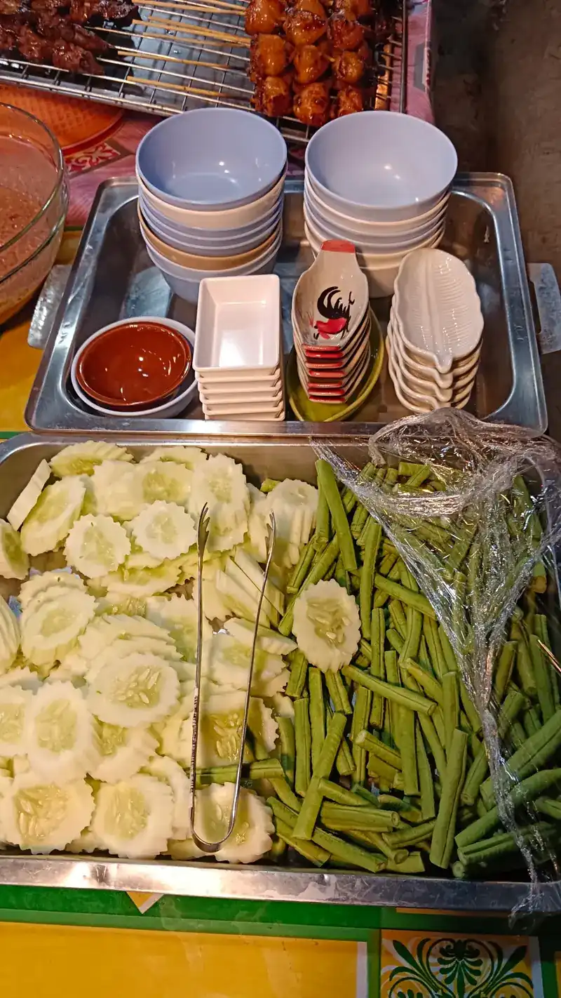 Trays of sliced cucumber and long beans, with skewers on the grill behind