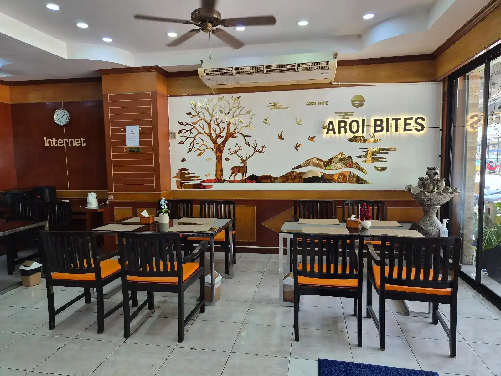 The dining room at Aroi Bites: dark wood chairs with orange cushions and the lit Aroi Bites sign on a white wall with a gold tree and deer / ห้องอาหารของอร่อยไบท์ เก้าอี้ไม้สีเข้มเบาะส้ม และป้ายไฟชื่อร้านบนผนังสีขาวลายต้นไม้และกวางสีทอง