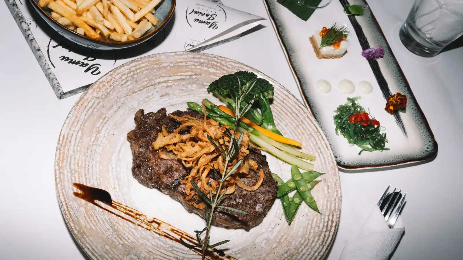 A steak with crispy onions, rosemary and greens, fries and small plates on a white table at Yamu Social