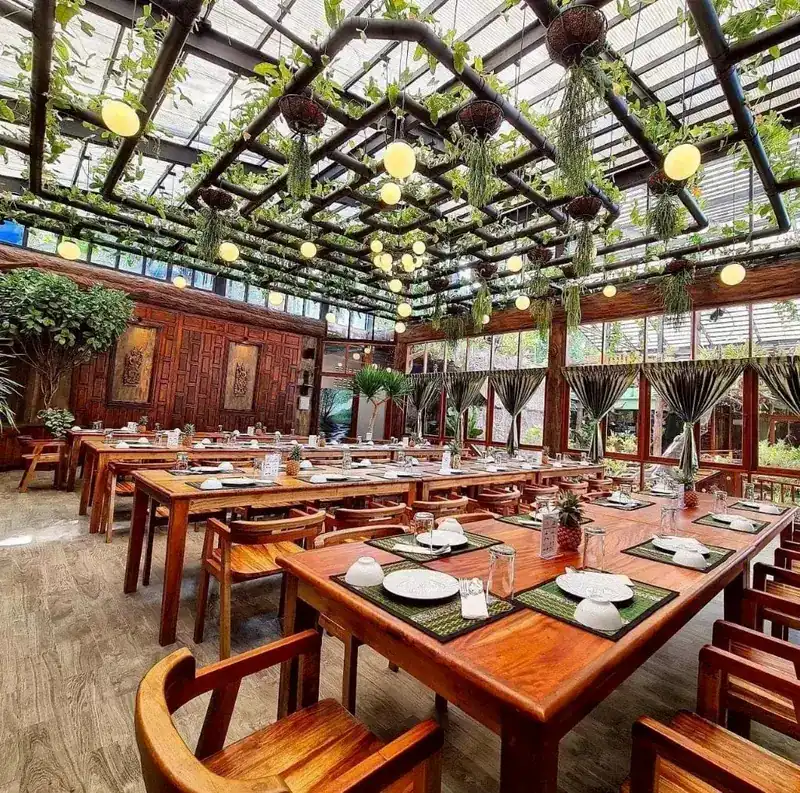 Dining room under a glass roof hung with plants and round lamps, long wooden tables set for dinner