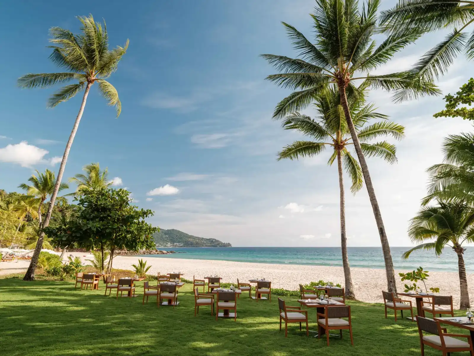 Dining tables on the lawn under tall coconut palms, with the white sand and turquoise sea of Pansea Beach behind