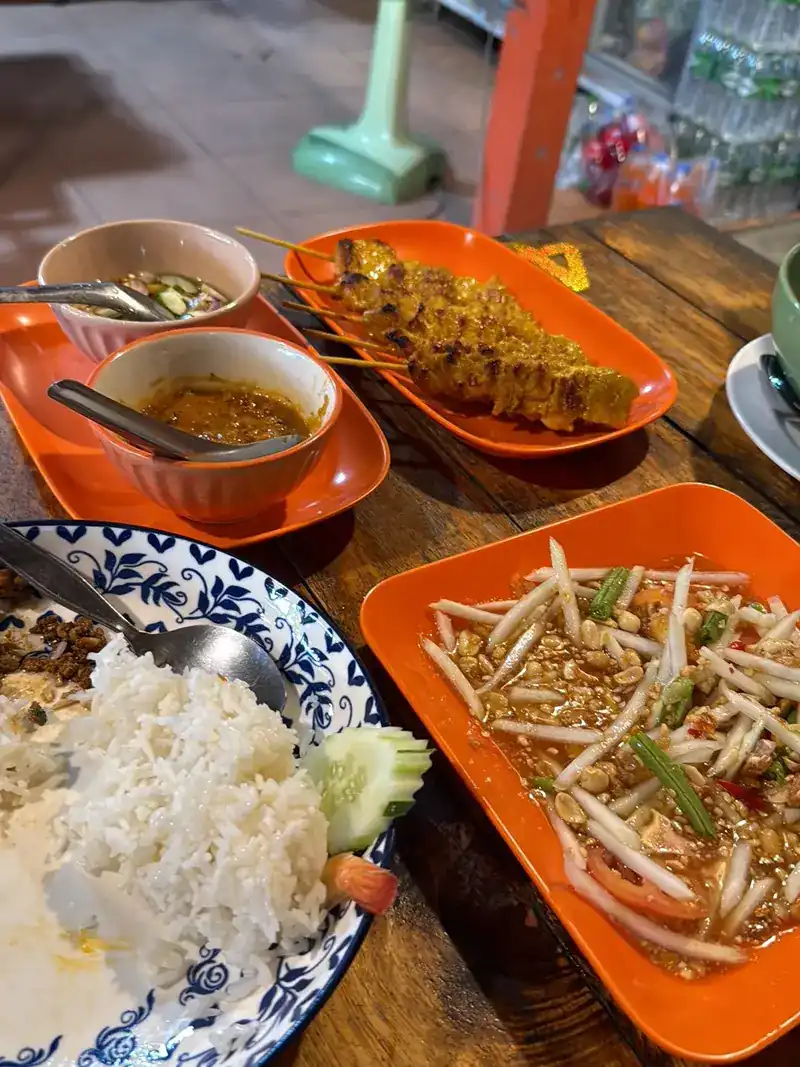 Papaya salad, satay with its sauce and a plate of rice on a wooden table / ส้มตำ สะเต๊ะกับน้ำจิ้ม และข้าวสวยบนโต๊ะไม้