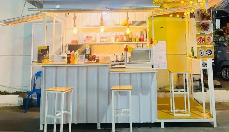 The kiosk at night: a white corrugated counter, tall stools, hanging bulbs and a yellow-lit kitchen