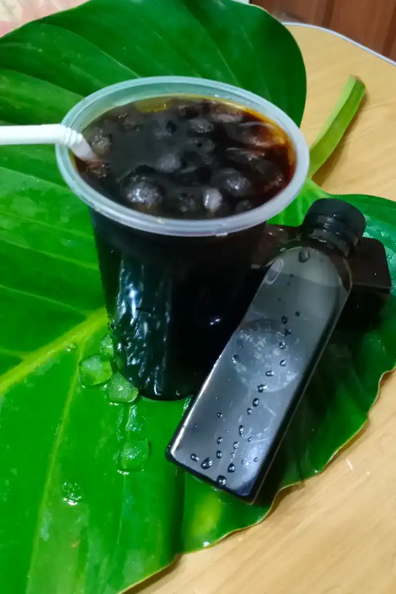 An iced dark drink in a cup with a bottle beside it, on a wet leaf