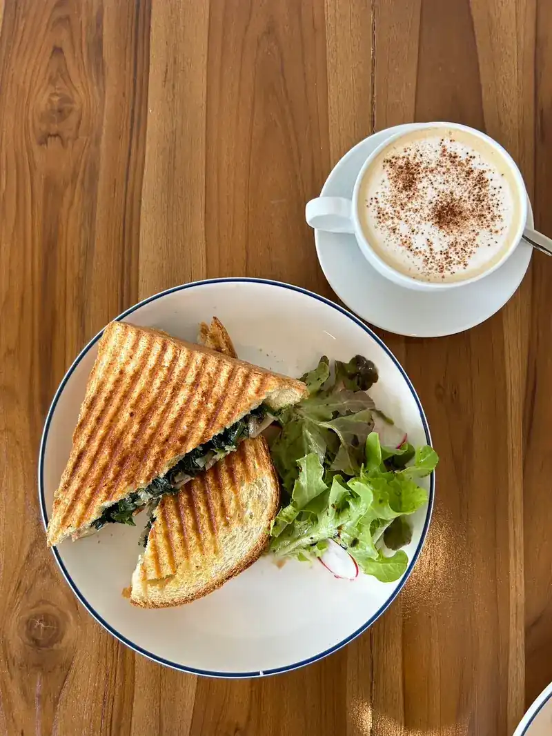 A grilled sandwich with salad on a white plate beside a cappuccino, seen from above on a wooden table