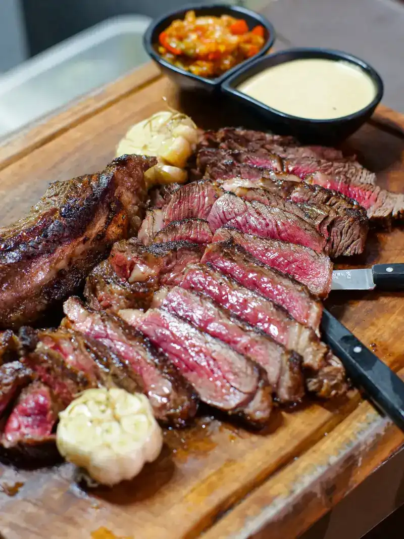 Grilled steak carved into thick slices on a wooden board, with roasted garlic and two sauces