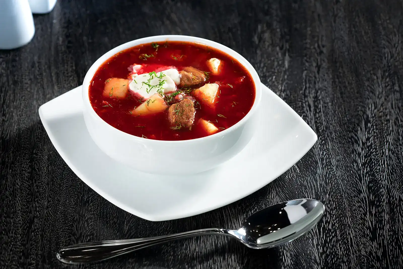 A white bowl of red borsh with beef, potato, sour cream and dill on a black wooden table