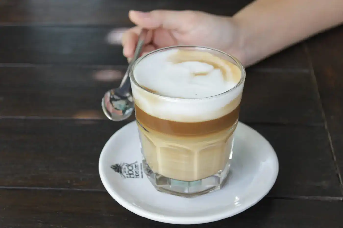 A hand stirring a layered milk coffee in a glass on a Hock Hoe Lee saucer, on a dark wooden table