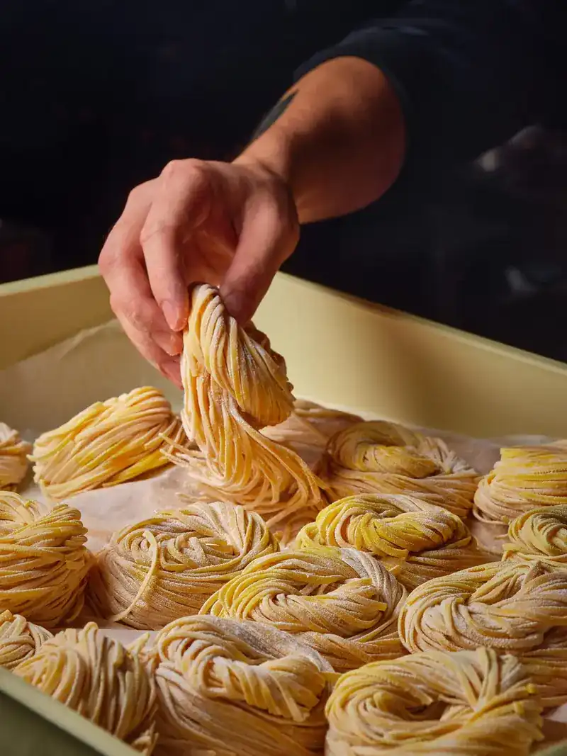 A hand lifting a nest of fresh pasta from a tray of pasta nests