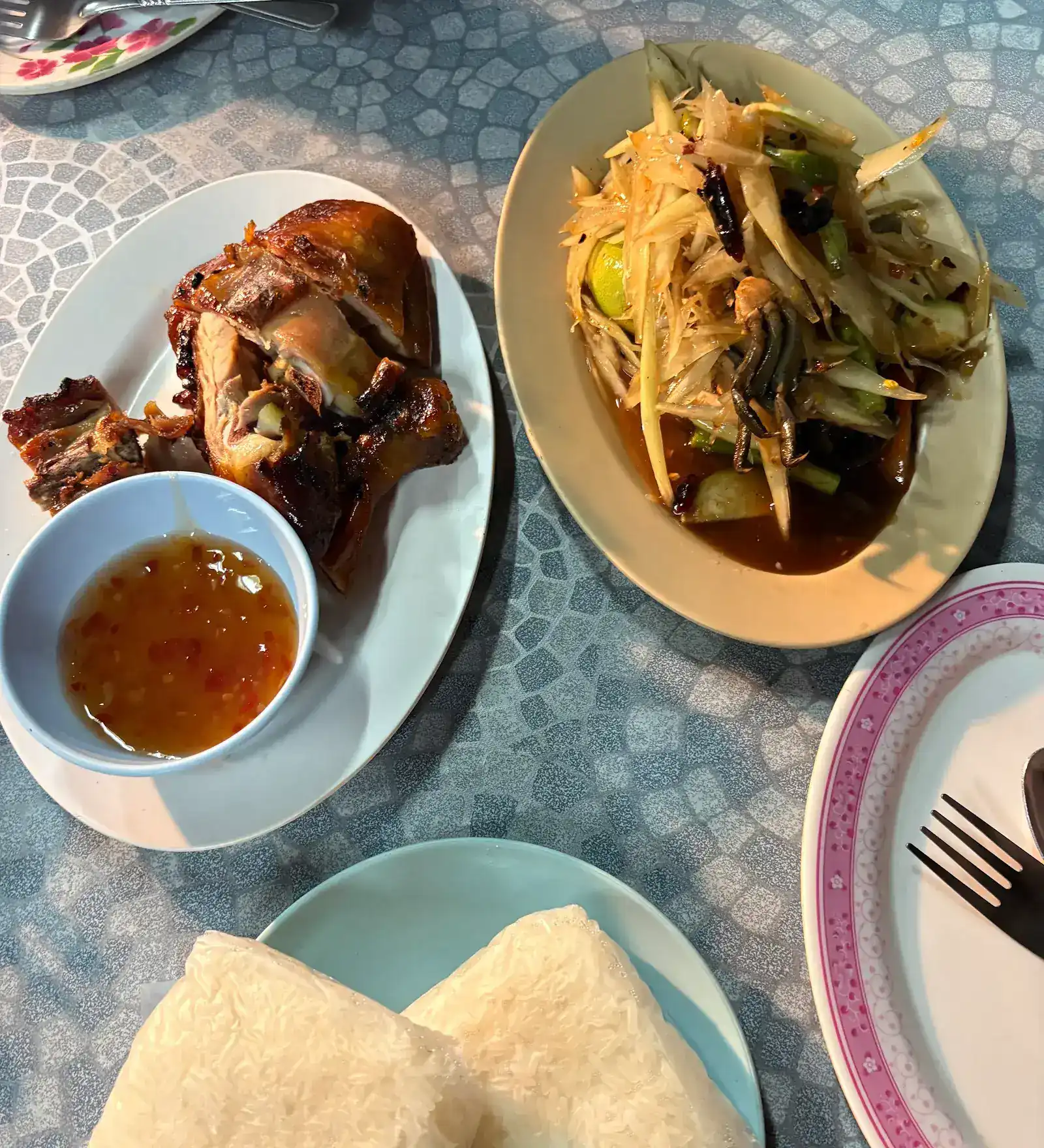 Grilled chicken with dipping sauce, papaya salad with crab and two parcels of sticky rice on a blue table