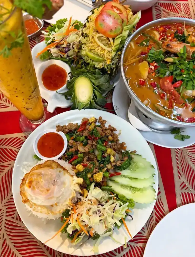 A table seen from above: pad kra pao with a fried egg, papaya salad, a pot of tom yum and a fruit smoothie