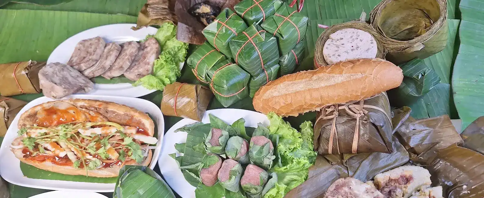 Vietnamese food laid out on banana leaves: a filled baguette, sliced pork rolls, parcels tied in banana leaf and wrapped nam on lettuce