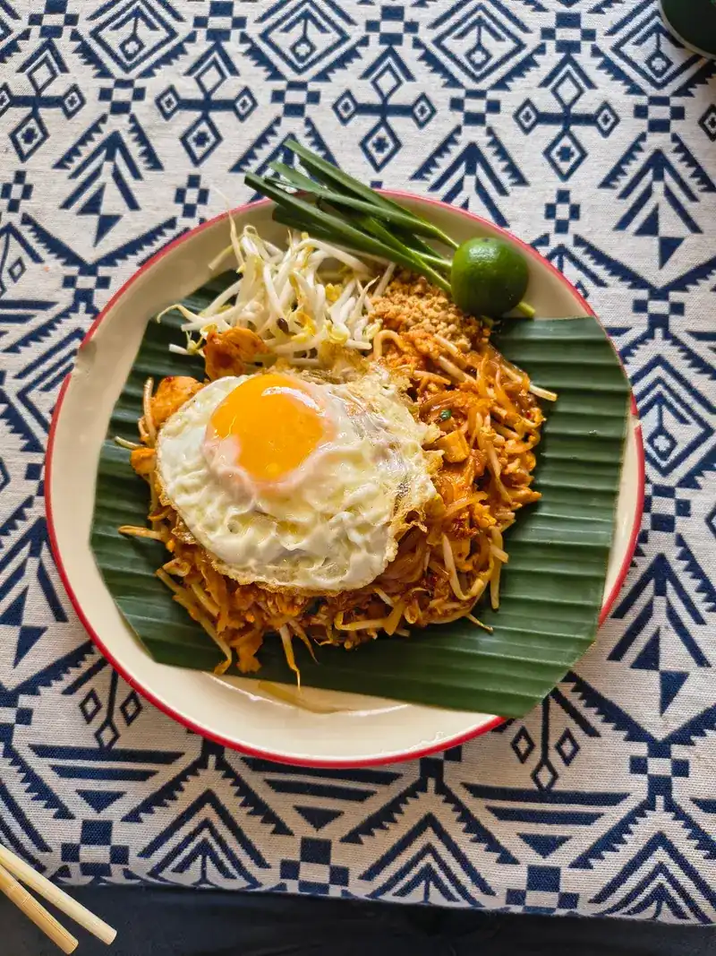 Pad thai topped with a fried egg on a banana leaf, on a blue and white woven cloth