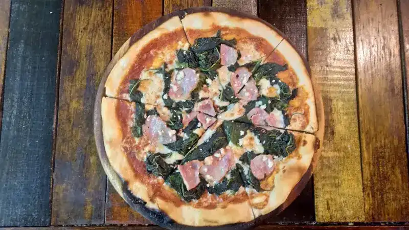 A thin-crust pizza with leafy greens and ham on a wooden board