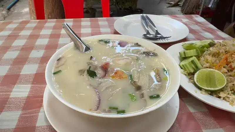 A bowl of coconut soup beside fried rice and lime on a checked tablecloth