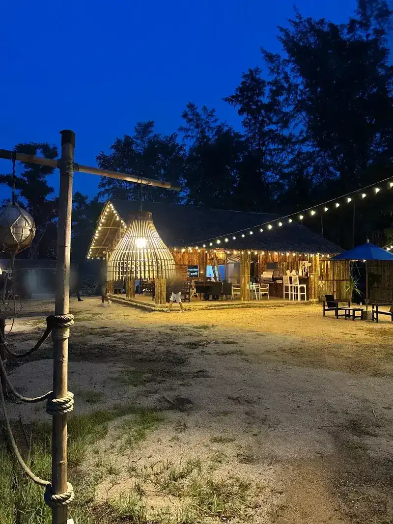 The thatched bamboo hall at dusk, with a large woven lamp, string lights and tables on the sand