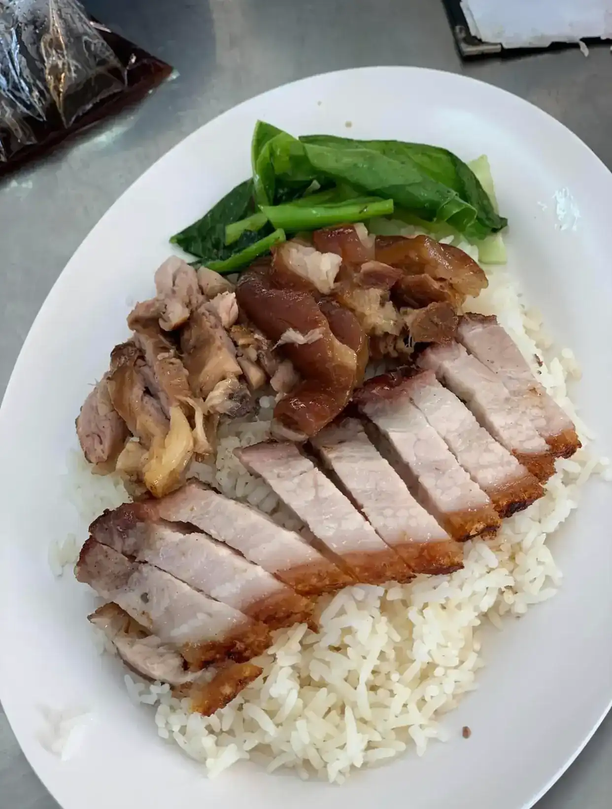 Sliced crispy pork belly and braised pork leg over white rice, with greens, on a white oval plate