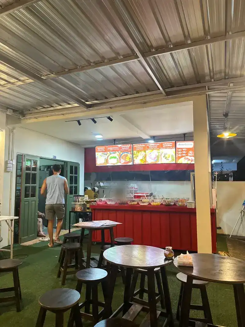 Seating under a metal roof: wooden stools on green turf, with the red counter behind
