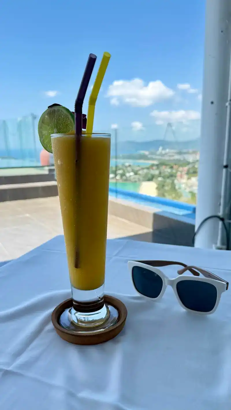 A yellow fruit smoothie and sunglasses on a white table, the sea behind / สมูทตี้ผลไม้สีเหลืองกับแว่นกันแดดบนโต๊ะสีขาว ทะเลอยู่ด้านหลัง