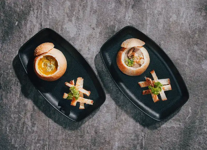 Two soups served in bread bowls with toast fingers on black plates