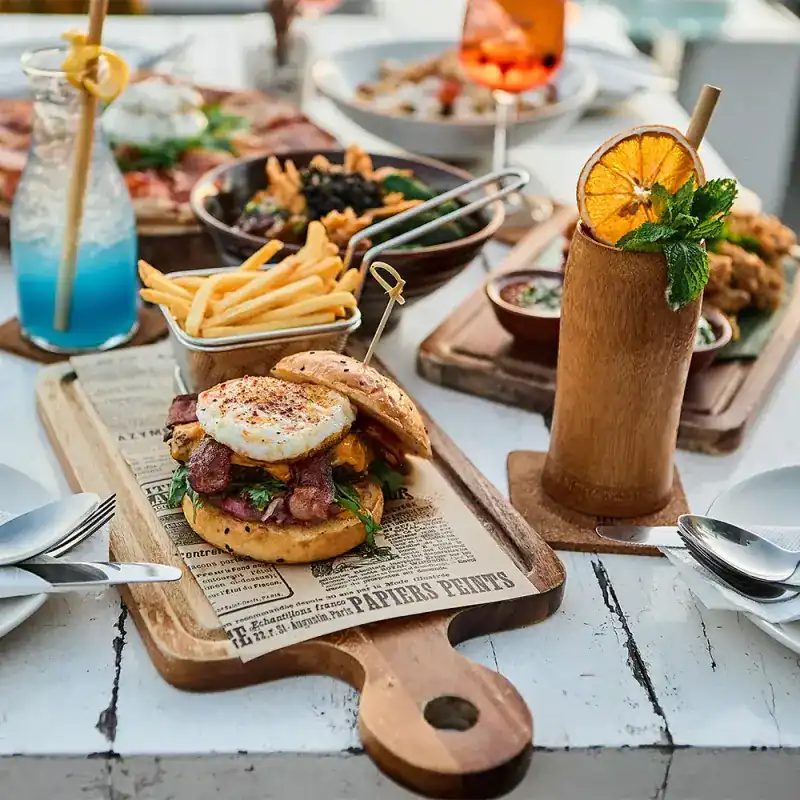 Burger with a fried egg and a basket of fries on a wooden board, a table set for sharing