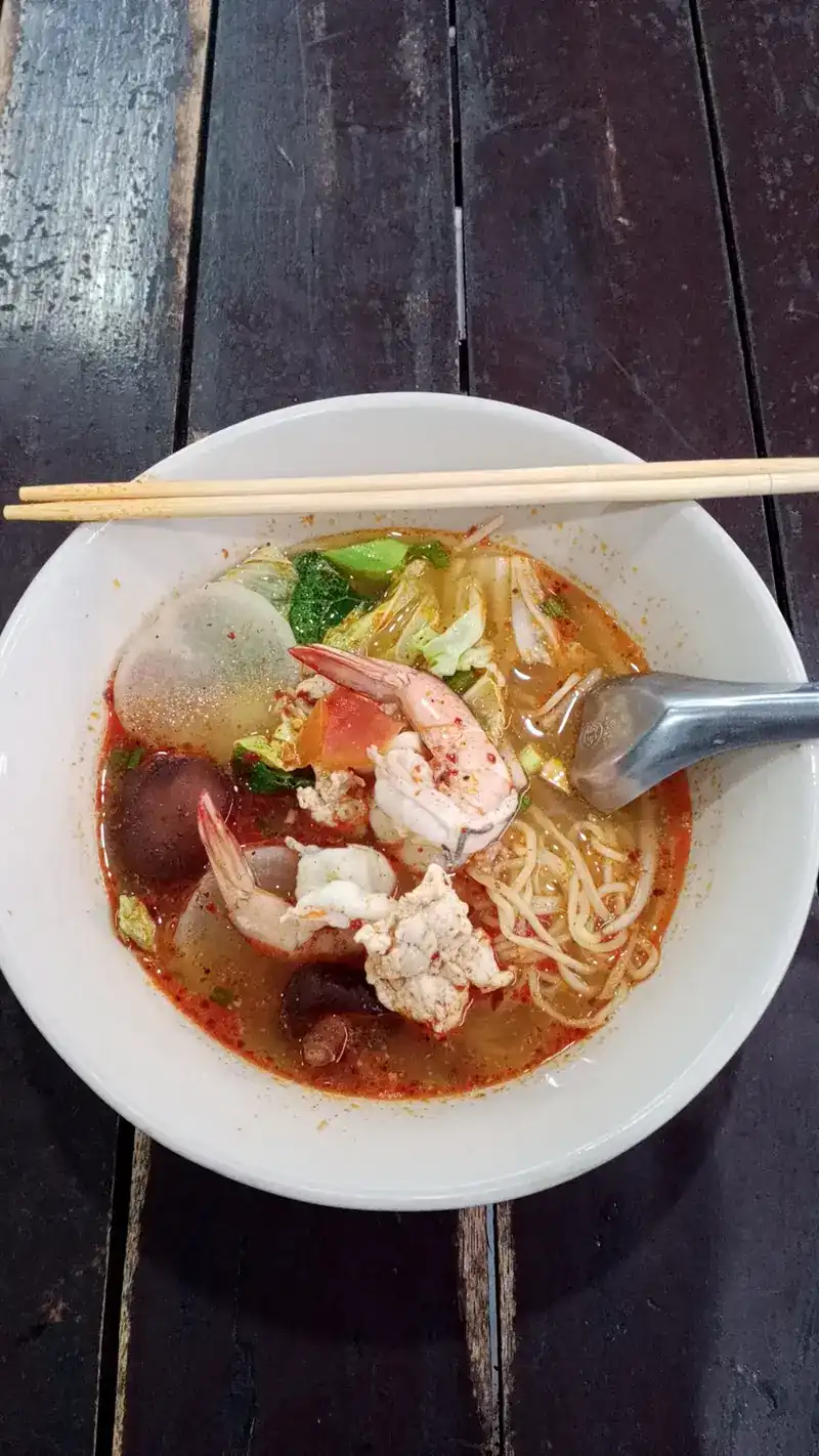 A bowl of red noodle soup with prawns on a wooden table