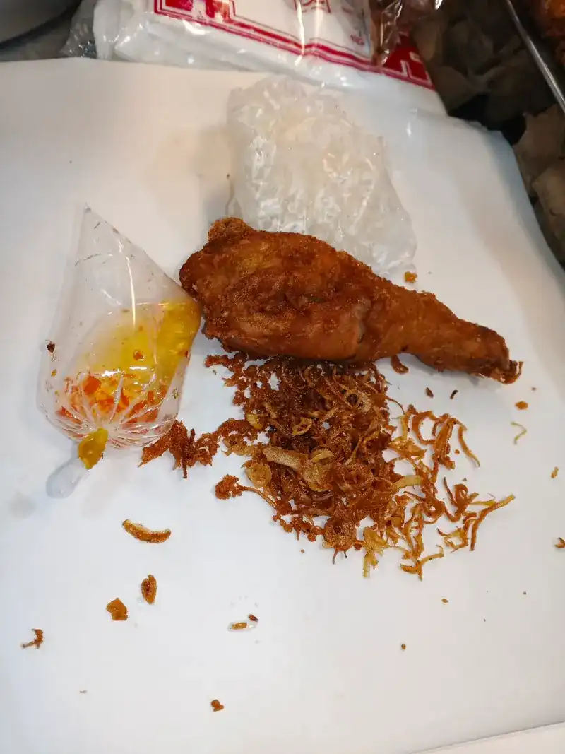 A piece of fried chicken with fried shallots and a bag of sauce