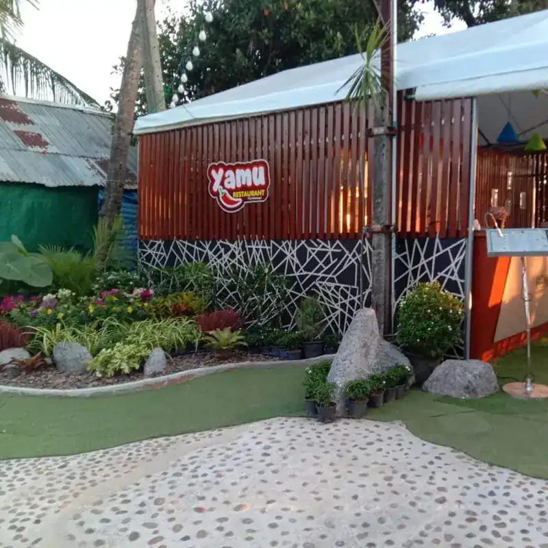 The entrance: a red Yamu sign on a timber wall, a flower garden and a pebble path