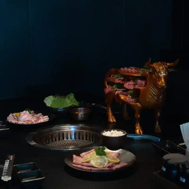 A dark table set for yakiniku: a grill sunk into the table, plates of sliced meat and the bronze bull stand