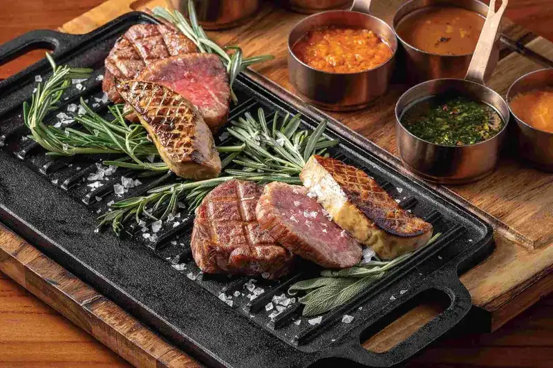 Grilled steak cuts on a cast-iron plate with rosemary, sea salt and copper pans of sauce
