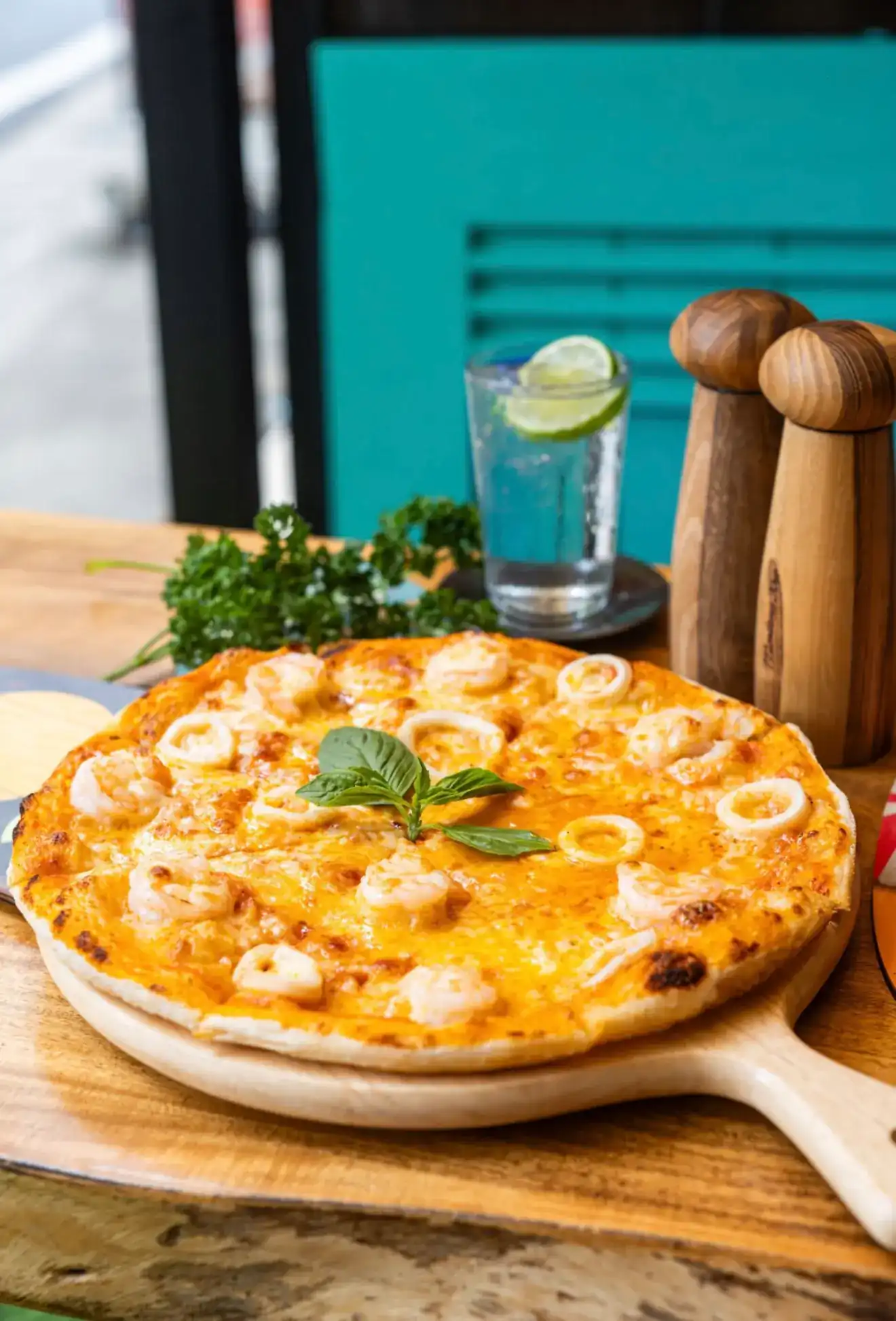 Seafood pizza with prawns and squid rings on a wooden board, a tall glass with lime behind it