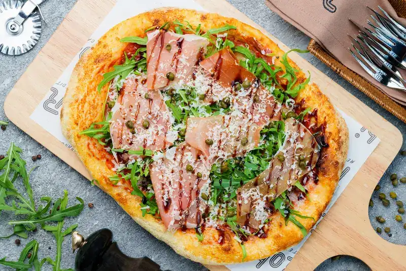 Oval Roman pizza topped with cured ham, rocket, capers and shaved cheese on a wooden board
