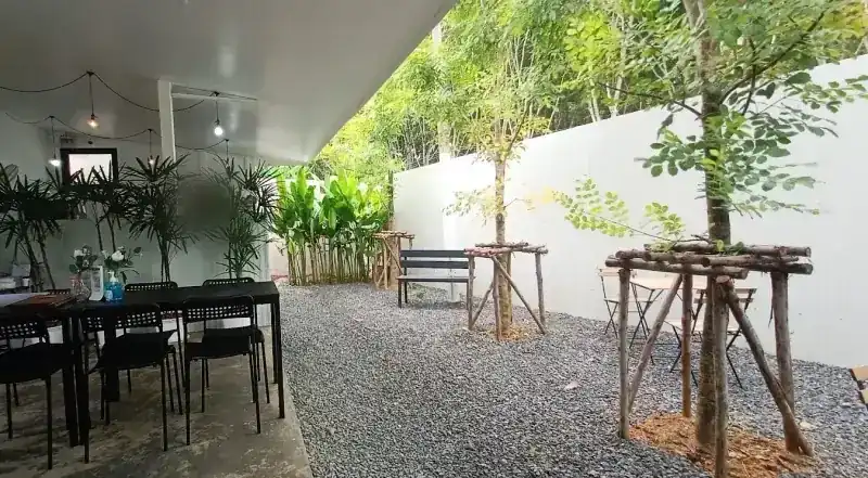 Long black tables under the roof and a gravel garden with young trees along a white wall