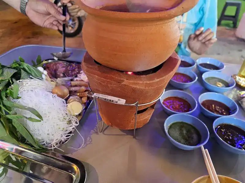 A clay pot on a charcoal stove, with rice noodles, fresh greens and small bowls of dipping sauce