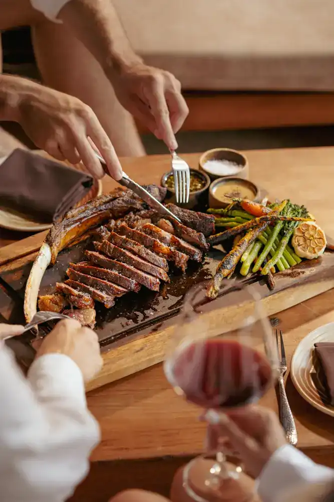 Hands carving a sliced steak on the bone, with grilled asparagus and carrots