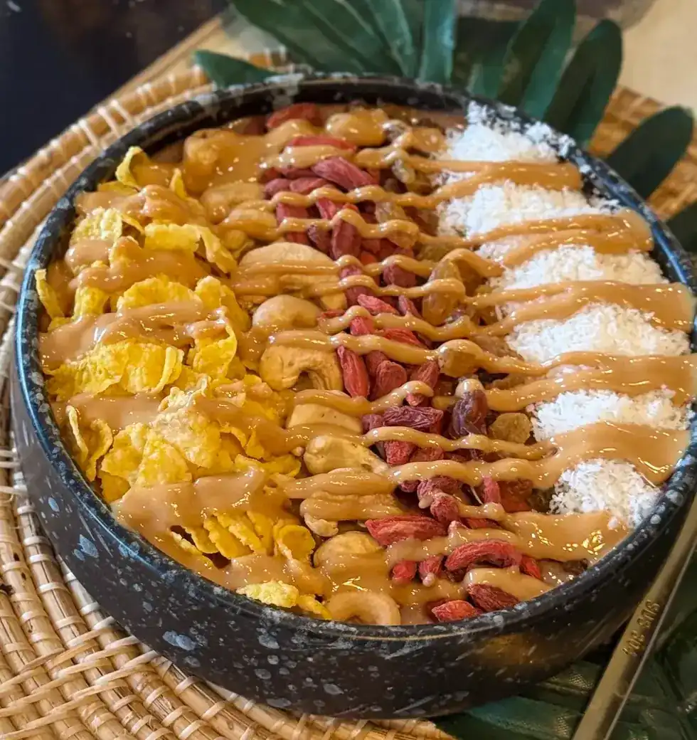 Açaí bowl topped with cornflakes, cashews, goji berries, coconut and peanut butter on a rattan mat